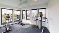 This fitness center offers a bright, contemporary workout environment designed to inspire an active lifestyle. The space is wrapped on multiple sides by large, black-framed floor-to-ceiling windows that flood the room with abundant natural light and provide expansive views of the surrounding urban landscape and tree canopy, creating an energizing atmosphere for exercise. The flooring throughout is fitted with dark rubber gym tiles, offering both durability and comfort underfoot. A ceiling fan overhead ensures air circulation during workouts. Equipment includes a NordicTrack elliptical machine, a stationary spin bike with an integrated display, and a NordicTrack functional trainer for strength and cable exercises. The clean, white walls contribute to an open and airy feel, complementing the unobstructed sightlines to the outdoors. Entry to the fitness center is through a glass-paneled door, alongside posted fitness center rules and a sanitizing station, emphasizing a clean and organized environment. The overall design balances functionality with a modern aesthetic, making this a well-appointed space suited for a comprehensive workout routine.