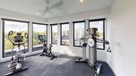 This fitness center offers a bright, contemporary workout environment designed to inspire an active lifestyle. The space is wrapped on multiple sides by large, black-framed floor-to-ceiling windows that flood the room with abundant natural light and provide expansive views of the surrounding urban landscape and tree canopy, creating an energizing atmosphere for exercise. The flooring throughout is fitted with dark rubber gym tiles, offering both durability and comfort underfoot. A ceiling fan overhead ensures air circulation during workouts. Equipment includes a NordicTrack elliptical machine, a stationary spin bike with an integrated display, and a NordicTrack functional trainer for strength and cable exercises. The clean, white walls contribute to an open and airy feel, complementing the unobstructed sightlines to the outdoors. Entry to the fitness center is through a glass-paneled door, alongside posted fitness center rules and a sanitizing station, emphasizing a clean and organized environment. The overall design balances functionality with a modern aesthetic, making this a well-appointed space suited for a comprehensive workout routine.
