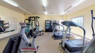 This fitness center is a fully outfitted, single-floor exercise facility designed to support a well-rounded workout routine. The space is furnished with a comprehensive selection of cardio equipment, including multiple treadmills, elliptical machines, and an upright stationary bike, all from Life Fitness. A multi-station cable weight machine and a rowing machine round out the strength and cross-training options. The room features light yellow walls, carpeted flooring, and bright fluorescent overhead lighting that keeps the space well-lit throughout. Large windows along one wall allow natural light to filter in and provide views of the surrounding exterior. A full-length wall mirror enhances the sense of openness and allows users to monitor their form during workouts. Practical amenities include a built-in water fountain, wall-mounted televisions for entertainment during cardio sessions, and beverage and snack vending machines. A wall clock, fire extinguisher, and clearly marked exit signage reflect the facility's attention to safety and functionality. The overall atmosphere is clean, organized, and purposefully equipped to meet the needs of regular gym-goers.