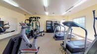 This fitness center is a fully outfitted, single-floor exercise facility designed to support a well-rounded workout routine. The space is furnished with a comprehensive selection of cardio equipment, including multiple treadmills, elliptical machines, and an upright stationary bike, all from Life Fitness. A multi-station cable weight machine and a rowing machine round out the strength and cross-training options. The room features light yellow walls, carpeted flooring, and bright fluorescent overhead lighting that keeps the space well-lit throughout. Large windows along one wall allow natural light to filter in and provide views of the surrounding exterior. A full-length wall mirror enhances the sense of openness and allows users to monitor their form during workouts. Practical amenities include a built-in water fountain, wall-mounted televisions for entertainment during cardio sessions, and beverage and snack vending machines. A wall clock, fire extinguisher, and clearly marked exit signage reflect the facility's attention to safety and functionality. The overall atmosphere is clean, organized, and purposefully equipped to meet the needs of regular gym-goers.