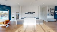 The Yardly Monroe sales center, developed by Taylor Morrison, is a thoughtfully designed single-floor welcome and information hub that offers a polished, professional environment for prospective residents. The open-plan layout is anchored by a sleek white reception desk positioned beneath a bold branded wall display, immediately establishing a clean and contemporary aesthetic upon entry.

Light hardwood flooring flows throughout the entire space, complementing the crisp white walls and bright recessed lighting overhead. A striking teal accent wall anchors the lounge area, where leather armchairs and a marble-top coffee table create a comfortable seating arrangement for consultations or casual conversations. Oversized windows along this wall frame views of an outdoor pool area, flooding the space with natural light.

Along the perimeter, dark gray cabinetry provides functional storage and a beverage station, while branded community maps and informational displays add character and context for visitors. A glass-paneled interior door leads to a private office space, offering a more intimate setting for detailed discussions. The entry area features floor-to-ceiling windows that bathe the open floor in generous sunlight, enhancing the airy and welcoming atmosphere. Decorative potted plants are strategically placed throughout, adding warmth and a touch of nature to the modern commercial interior.