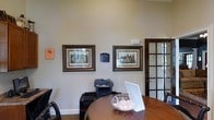 This single-floor professional office space combines functionality with a polished, classic aesthetic. The reception or meeting area centers around a warm, rich wood table, complemented by traditional dark-framed seating that adds to the space's refined character. Neutral, soft beige walls provide a calm backdrop throughout, enhancing the sense of openness and light.

The walls are adorned with elegantly framed architectural prints featuring detailed illustrations of historic buildings, lending the space a distinguished and cultured ambiance. A small placard displayed between the two prints adds an informational or branding element, reinforcing the professional identity of the space.

Practical office equipment, including a multifunction printer, is neatly integrated into the workspace without detracting from the overall aesthetic. A white display stand on the table suggests an interactive or informational component for visitors.

A classic wood-framed glass-paned door on the right side of the space adds architectural character and allows for a sense of visual connectivity between areas. Neutral carpeted flooring ties the room together cohesively. The overall ambiance is professional, tidy, and thoughtfully appointed — well-suited for client-facing interactions or administrative functions.