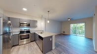 Located at 492 E Washington St in North Attleboro, Massachusetts, this single-floor residence offers a thoughtfully designed open-concept layout that seamlessly connects the kitchen, dining, and living areas. The kitchen is a standout feature, equipped with full stainless steel appliances including a range, microwave, dishwasher, and refrigerator. Sleek gray cabinetry pairs with granite-style countertops and a peninsula island accented by pendant lighting, providing both functional workspace and a casual dining option. Light wood-style flooring runs throughout the main living areas, contributing to a clean, contemporary aesthetic. The living area benefits from a sliding glass door that invites natural light and offers outdoor views, complemented by a ceiling fan for year-round comfort. The bedroom features neutral-toned carpet, large windows with outdoor views, and ample wall space. The bathroom is well-appointed with a granite-topped vanity, a tub-shower combination, and matching light wood flooring that ties the space together cohesively. The entryway provides a practical corridor connecting all areas of the unit, creating an efficient and welcoming flow throughout this modern residence.
