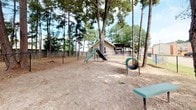 This outdoor community space is thoughtfully designed to offer both functionality and a welcoming natural atmosphere. At its heart is a spacious, fully enclosed dog park surrounded by chain-link fencing, providing a safe and open environment for pets to roam freely. The park is shaded by several mature trees, including a prominent large-canopy tree at its center, which offers natural relief from the sun. A variety of agility and play equipment is distributed throughout the park, including ramps, a tunnel, and a tire swing, catering to active pets. Seating benches are positioned around the perimeter for convenience. Adjacent to the dog park, a covered boardwalk-style walkway runs along the exterior of a single-story building with light-colored lap siding and numbered units. The walkway features a latticed pergola overhead, decorative cast-iron planters, and wooden railings, giving it a charming, cottage-like character. Landscaped beds with decorative river rock, succulents, and ornamental plants border the building's exterior. The broader grounds include well-maintained lawns, additional mature trees such as magnolias, and ample parking, all set against a backdrop of lush wooded surroundings.