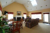 Located at 3245 Meadows Way in Chesapeake, Virginia, this single-floor residence offers a well-appointed layout that balances comfort and functionality. The centerpiece of the home is the expansive living and dining area, distinguished by soaring vaulted ceilings that create a bright, airy atmosphere. Skylights positioned along the roofline flood the space with natural light, enhancing the warm, neutral tones of the carpeted floors and painted walls. A classic white-mantled fireplace with a marble surround serves as a striking focal point, flanked by armchairs and complemented by a striped sofa arrangement ideal for entertaining. Large windows dressed with valances and blinds allow for controlled natural light while maintaining a polished aesthetic. A dedicated dining area with direct access to the outdoors provides additional versatility. The kitchen is thoughtfully designed with light wood cabinetry extending to the ceiling, white appliances including a built-in microwave and dishwasher, generous counter space, and hardwood flooring accented by a decorative area rug. Cabinetry storage is abundant, with upper cabinets reaching toward the ceiling for maximum utility. Overall, the space presents a traditional, comfortable character well-suited for everyday living.