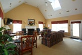 Located at 3245 Meadows Way in Chesapeake, Virginia, this single-floor residence offers a well-appointed layout that balances comfort and functionality. The centerpiece of the home is the expansive living and dining area, distinguished by soaring vaulted ceilings that create a bright, airy atmosphere. Skylights positioned along the roofline flood the space with natural light, enhancing the warm, neutral tones of the carpeted floors and painted walls. A classic white-mantled fireplace with a marble surround serves as a striking focal point, flanked by armchairs and complemented by a striped sofa arrangement ideal for entertaining. Large windows dressed with valances and blinds allow for controlled natural light while maintaining a polished aesthetic. A dedicated dining area with direct access to the outdoors provides additional versatility. The kitchen is thoughtfully designed with light wood cabinetry extending to the ceiling, white appliances including a built-in microwave and dishwasher, generous counter space, and hardwood flooring accented by a decorative area rug. Cabinetry storage is abundant, with upper cabinets reaching toward the ceiling for maximum utility. Overall, the space presents a traditional, comfortable character well-suited for everyday living.