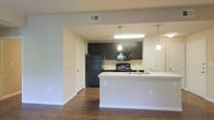 This single-floor space is designed with an open-concept layout that promotes a seamless flow between the kitchen and living areas. The kitchen serves as a central highlight, featuring dark espresso-toned cabinetry that contrasts sharply against light-colored countertops and neutral grey walls. Black appliances, including a full-size refrigerator, electric range, and microwave, lend a sleek, cohesive aesthetic to the space. A peninsula island with an integrated sink extends into the main living area, providing both additional prep space and a natural gathering point. Pendant lighting fixtures hang above the peninsula, adding focused illumination and a touch of contemporary style. The living area is finished with rich, dark hardwood-style flooring that runs throughout, grounding the space with warmth and depth. Recessed ventilation and flush ceiling lighting contribute to a clean, uncluttered overhead aesthetic. White trim and baseboards frame the neutral grey walls, providing crisp definition throughout. Multiple doorways offer convenient access to additional rooms, enhancing the overall functionality of the layout. The overall ambiance is modern and practical, with a neutral palette that provides a versatile backdrop for a variety of interior styles.
