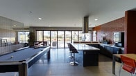 This contemporary amenity lounge is designed with both style and function in mind, offering an open-plan layout that seamlessly blends social and recreational spaces. The centerpiece of the room is a large kitchen island topped with a light-colored countertop and lined with modern bar-height stools, complemented by a professional-grade overhead range hood and sleek dark cabinetry along the perimeter. A full billiards table anchors the lounge area, providing a focal point for entertainment. The space features large-format tile flooring in a cool grey tone, lending a polished and cohesive feel throughout. Recessed lighting keeps the interior bright and well-illuminated, while pendant lights add a layer of ambient warmth above the kitchen zone. An accent wall clad in exposed brick introduces a warm, industrial contrast to the otherwise modern palette. Floor-to-ceiling sliding glass doors span the rear wall, flooding the space with abundant natural light and offering direct access to an outdoor patio area, where additional seating and landscaping are visible. The overall ambiance is sophisticated and inviting, making it well-suited as a shared gathering and entertainment space.