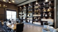 Located at 740 Bridgeport Ave in Shelton, Connecticut, this single-floor space presents a refined and sophisticated interior designed with a strong attention to detail. The standout feature of the space is a dramatic floor-to-ceiling built-in shelving unit finished in deep, dark tones, elegantly illuminated by individual spotlights that highlight curated décor pieces, books, sculptural objects, and decorative accents. The shelving serves as both a functional and artistic focal point, lending the space a library-like grandeur. Natural light filters in through windows, complementing the warm ambient glow from the built-in lighting. The flooring features wide-plank hardwood in a light, natural finish, creating a balanced contrast against the dark shelving and rich wall tones. A substantial wooden work surface paired with executive-style seating anchors the room, reinforcing its professional character. The overall ambiance is polished and upscale, blending modern design sensibilities with classic, timeless elements. The thoughtful layering of lighting, materials, and curated décor creates an environment that feels both highly functional and visually impressive — ideal for professional or creative use.