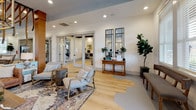 Located at 4140 Bayswater Rd in Winterville, NC, this thoughtfully designed single-floor amenity space combines modern aesthetics with warm, welcoming interiors throughout. The main lounge area features light hardwood flooring, recessed lighting, and a sophisticated statement chandelier with a geometric brass frame. Comfortable seating arrangements are complemented by French glass doors that open to an outdoor patio, flooding the space with natural light and offering views of the surrounding community. A dedicated café-style dining area serves as a standout feature, with a teal upholstered banquette, butcher block tables, white bistro chairs, and a dramatic ceiling installation of hanging rope pendants with globe lights — lending the space a coastal, resort-like ambiance. The catering kitchen is fully appointed with soft gray shaker cabinetry, white quartz countertops, a textured subway tile backsplash, stainless steel appliances including double wall ovens and a side-by-side refrigerator, open display shelving, and a utility sink — ideal for hosting events. A welcoming reception area with wood-look flooring, paired wall mirrors, and glass-paned interior doors leads to additional private rooms, including what appears to be a fitness area. The overall design palette of soft whites, warm wood tones, and brushed gold accents creates a cohesive, upscale atmosphere throughout.