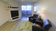 This single-floor space offers a clean, modern layout designed for comfortable everyday living. The living area serves as the heart of the space, showcasing smooth hardwood-style flooring that runs throughout and lends a warm, cohesive feel to the interior. A sleek white fireplace with a minimalist mantle serves as a striking focal point, adding both style and function to the room. Large vertical blinds frame a expansive sliding glass door that floods the space with natural light and opens directly onto a private balcony, offering a seamless connection between indoor and outdoor living. The living area is well-proportioned, accommodating seating comfortably while maintaining an open, airy atmosphere. Neutral wall tones complement the dark leather furnishings and a textured area rug, creating a balanced contrast throughout. The overall ambiance is calm and contemporary, with thoughtful design details that enhance both the visual appeal and practicality of the space. The combination of natural light, a functional fireplace, and balcony access makes this a versatile and inviting environment suited for relaxing or entertaining.
