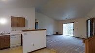This single-floor space is defined by its vaulted ceilings, which lend a sense of height and openness throughout the main living area. The open-concept layout connects the living space to an adjacent area divided by a half-wall with a wood-trimmed ledge, providing subtle separation while maintaining visual flow between spaces. Upper cabinetry visible beyond the half-wall suggests a kitchen or utility area nearby, with a doorway leading further into the unit.

The living area is finished with wall-to-wall carpet in a neutral gray tone, complementing the clean white walls and dark wood baseboards throughout. A sliding glass door at the far end of the living space serves as both a source of natural light and a connection to the outdoors, flooding the room with daylight and offering views beyond. A floor vent near the sliding door indicates a climate control system integrated into the flooring.

The overall ambiance is clean, neutral, and versatile, with the vaulted ceiling and open layout making the space feel larger than its footprint suggests. The design provides a functional and adaptable environment suitable for a variety of furnishing arrangements.