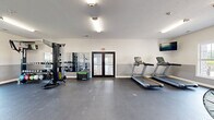 Located at 5504 Town Center Dr in Mishawaka, Indiana, this fitness center offers a well-equipped, single-floor exercise space designed to support a wide range of workout routines. The room features a clean, modern aesthetic with two-tone gray and white walls, durable rubber flooring, and bright overhead lighting that keeps the environment energized and functional.

Natural light filters in through multiple windows along the perimeter walls, offering pleasant outdoor views and a welcoming atmosphere. The cardio section is well-stocked with multiple Matrix treadmills, an air bike, and a rowing machine, complemented by a wall-mounted flat-screen television for entertainment during workouts. A strength training area features a power rack with weight plates and a dedicated functional training station — a STACK unit complete with a full dumbbell rack, cable attachments, and battle ropes for high-intensity training.

A large wall mirror on one side of the room enhances the sense of space and supports form-checking during exercises. Additional equipment includes a plyometric box for jump training and a weight storage area to keep the floor organized. Access is provided through both a set of glass double doors and a secondary door, ensuring convenient entry and exit. The space is thoughtfully arranged to provide ample open floor area for stretching, functional movements, and free-form training.