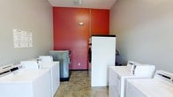 This single-floor laundry room is designed for shared use, offering a practical and efficient layout equipped with multiple top-loading and front-loading washers and dryers. The room's most distinctive design element is its bold red-orange accent wall, which contrasts with the neutral grey-beige walls on either side, adding a lively visual focal point to an otherwise utilitarian space. The machines are arranged along the perimeter, maximizing the usable floor area and allowing for comfortable movement between units. A dark-topped surface is positioned between machines, providing a convenient area for sorting or folding laundry. The flooring consists of durable tile in a neutral tan tone, well-suited to the demands of a high-use laundry facility. Overhead lighting keeps the room well-illuminated. A vertical pipe runs along the accent wall, indicative of the room's functional infrastructure. Overall, the space is straightforward and purpose-driven, prioritizing accessibility and capacity while incorporating a touch of color to enliven the environment.