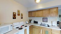 This two-floor model home offers a well-organized and functional layout designed to maximize comfort and everyday livability. The kitchen serves as a central highlight, featuring warm honey-toned oak cabinetry that lines both the upper and lower walls, providing generous storage space for all culinary needs. White laminate countertops offer a clean, bright contrast to the natural wood tones, while a stainless steel sink adds a practical touch. A white coil-burner range anchors one end of the kitchen, making the space fully equipped for home cooking. The cabinetry layout creates an efficient L-shaped workspace that flows naturally through the room. Adjacent to the kitchen, louvered bifold closet doors hint at additional storage solutions thoughtfully integrated into the floor plan. The flooring transitions from dark hardwood in the kitchen to carpet in the connecting areas, adding warmth and variety throughout the home. Neutral wall tones in soft white and sage green contribute to a calm, welcoming ambiance across both levels. With its practical design, ample storage, and cohesive aesthetic, this two-floor model home presents a comfortable and move-in-ready living environment suited to a variety of lifestyles.
