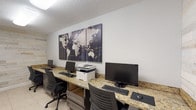 This single-room business center is designed to meet the productivity needs of its users with a clean, modern aesthetic. A continuous granite-topped workstation runs along the perimeter of the room, providing ample desk space for multiple users simultaneously. Each station is equipped with a desktop computer, keyboard, and mouse, while a shared multifunction printer and scanner sits conveniently within reach at the center of the counter. Ergonomic task chairs in a dark finish complement the warm tones of the granite surface and the charcoal cabinetry below, which offers built-in storage. The room is finished with light-toned ceramic tile flooring that keeps the space feeling bright and clean. A distinctive reclaimed wood plank accent wall adds a textural, rustic contrast to the otherwise contemporary interior, creating visual interest and warmth. A triptych world map canvas serves as the room's focal point, reinforcing the space's global, connected theme. Overhead flush-mount lighting ensures consistent illumination throughout, and a window along one wall allows for a degree of natural light. The overall ambiance is functional yet thoughtfully designed, making it a practical and comfortable workspace.