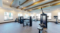 Located at 22274 E Quincy Ave in Aurora, CO, this fitness center offers a generously sized, single-floor workout facility designed to accommodate a comprehensive range of exercise needs. The space is defined by its striking architectural character, most notably the exposed wood ceiling beams that run the length of the room, adding a warm, rustic contrast to the otherwise clean, modern aesthetic. Ceiling fans and recessed lighting are integrated throughout, ensuring the space remains well-lit and comfortable during workouts.

The facility is divided into distinct functional zones. The cardio area lines one side of the room and features a row of treadmills, stationary bikes, and elliptical machines from brands including Spirit and True. The strength training section is equally well-appointed, housing a power rack with plate storage, a full dumbbell rack, cable machines, and a variety of selectorized resistance machines. Large wall mirrors in the strength area enhance the sense of space and allow for proper form checks during exercise.

Neutral-toned carpet tiles cover the floors throughout, providing comfort underfoot while maintaining a clean, professional appearance. Multiple windows along the walls allow natural light to filter in, offering views of the surrounding outdoor area. A storage unit and monitor station are also present, adding functional utility to the space. The overall ambiance is modern, organized, and purpose-built for fitness.