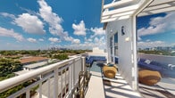 Located at 1640 East Sunrise Boulevard in Fort Lauderdale, Florida, this outdoor balcony offers a striking elevated vantage point with panoramic views of the Fort Lauderdale cityscape and surrounding lush tropical canopy. The balcony is finished in crisp white stucco, consistent with the building's clean, contemporary coastal architectural aesthetic. A white railed perimeter frames the open-air space, allowing unobstructed sightlines across the city skyline and expansive South Florida sky. The balcony is appointed with a coordinated outdoor seating arrangement, featuring a cushioned blue sectional sofa, a sleek round coffee table, and a woven accent chair in warm gold tones, all set atop a patterned area rug. A wall-mounted exterior light fixture provides illumination for evening use. The overhead pergola-style canopy offers partial shade while preserving the open, airy character of the space. Bathed in abundant natural sunlight, this balcony captures the essence of Florida outdoor living, serving as an ideal retreat to enjoy the vibrant energy of Fort Lauderdale from a comfortable, well-appointed perch high above the surrounding neighborhood.