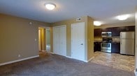 This single-floor residential unit offers a practical and comfortable layout designed for everyday living. The main living area features neutral-toned carpet flooring and warm beige walls, creating a welcoming atmosphere. A textured ceiling with flush-mount lighting adds a classic touch throughout the space. A set of bifold closet doors provides generous storage directly off the living area, complemented by an adjacent single-panel door leading to a separate room or utility space. The kitchen, visible from the main living area, is outfitted with dark espresso cabinetry, stone-look countertops, stainless steel appliances including a range and over-the-range microwave, and durable tile flooring — offering a functional and stylish cooking environment. A hallway visible from the living area connects to additional rooms, suggesting a well-organized floor plan. HVAC vents are integrated into the walls, ensuring year-round climate control. White trim and baseboards throughout provide clean contrast against the warm wall tones, contributing to a cohesive and tidy interior aesthetic. Overall, the space balances functionality with comfort in a straightforward, move-in-ready design.