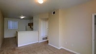 This single-floor space features an open layout that connects the living area to the kitchen, creating a functional and cohesive floor plan. The living area is finished with wall-to-wall carpeting in a neutral beige tone, complemented by light-colored walls and white baseboards that contribute to a clean, understated aesthetic. A flush-mount ceiling light provides overhead illumination for the main living space.

The kitchen is positioned adjacent to the living area and is accessible via a raised counter or pass-through bar, which adds a sense of openness while maintaining a defined separation between spaces. The kitchen features medium-toned wooden cabinetry, visible appliances including a refrigerator and microwave, and overhead fluorescent lighting. A white paneled door, likely leading to a closet or utility space, is recessed into a nook near the living area and sits above a floor-level vent, suggesting a dedicated HVAC intake. Additional vents are positioned near the ceiling, indicating a central air system throughout. The overall ambiance is simple, practical, and move-in ready.