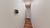 This efficiently designed 2-bedroom, 1-bathroom single-floor unit offers a well-organized layout that maximizes its compact footprint. Warm hardwood-style flooring runs consistently throughout the space, creating a cohesive and inviting atmosphere across all rooms.

The kitchen is a functional highlight, featuring gray shaker-style cabinetry, light granite-look countertops, and stainless steel appliances including a refrigerator and electric range. The L-shaped layout provides ample counter and storage space, and the kitchen opens to the main living area for an accessible flow.

A central hallway connects the main living spaces to the bedrooms and bathroom, and is equipped with a wall-mounted smart panel. The hallway leads to a louvered-door closet, adding practical storage to the unit.

The primary bedroom is a well-proportioned room featuring multiple windows that allow natural light to filter in, along with two panel doors offering access to a closet. The second bedroom is more intimate in scale, with a single window and the same warm hardwood-style flooring.

The bathroom is clean and simply appointed with neutral tile flooring, a pedestal-style vanity, toilet, and a mirrored medicine cabinet — providing all essential amenities in a tidy, efficient space.