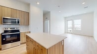 This contemporary single-floor unit showcases a clean, modern design with light wood flooring flowing throughout the open-concept layout. The well-appointed kitchen serves as the heart of the space, featuring flat-panel wood-tone cabinetry that extends to the ceiling for generous storage, a white quartz countertop, and a tiled backsplash in a soft neutral palette. A spacious central island provides additional prep space and casual seating potential. Stainless steel appliances — including a full-size refrigerator, electric range, over-the-range microwave, and dishwasher — complete the kitchen's functional and polished look. The kitchen opens seamlessly into the living area, which is bathed in natural light from large windows and a glass-panel door leading to a private balcony with outdoor views. The bedroom offers a bright, airy atmosphere with light-colored walls and natural light from a tall narrow window. The bathroom features large-format neutral tile in the tub surround with a deep soaking tub and modern chrome fixtures. A dedicated in-unit laundry closet with a stacked washer and dryer adds practical convenience. The overall ambiance is bright, fresh, and minimalist, with a cohesive neutral color palette throughout.
