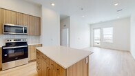 This contemporary single-floor unit showcases a clean, modern design with light wood flooring flowing throughout the open-concept layout. The well-appointed kitchen serves as the heart of the space, featuring flat-panel wood-tone cabinetry that extends to the ceiling for generous storage, a white quartz countertop, and a tiled backsplash in a soft neutral palette. A spacious central island provides additional prep space and casual seating potential. Stainless steel appliances — including a full-size refrigerator, electric range, over-the-range microwave, and dishwasher — complete the kitchen's functional and polished look. The kitchen opens seamlessly into the living area, which is bathed in natural light from large windows and a glass-panel door leading to a private balcony with outdoor views. The bedroom offers a bright, airy atmosphere with light-colored walls and natural light from a tall narrow window. The bathroom features large-format neutral tile in the tub surround with a deep soaking tub and modern chrome fixtures. A dedicated in-unit laundry closet with a stacked washer and dryer adds practical convenience. The overall ambiance is bright, fresh, and minimalist, with a cohesive neutral color palette throughout.