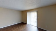 This single-floor space presents a straightforward and functional design. The main living area is finished with rich, dark hardwood-style flooring that contrasts cleanly against the warm beige walls, creating a welcoming atmosphere. A textured popcorn ceiling adds a classic touch overhead, while white baseboards provide a crisp border throughout the room.

A prominent sliding glass door serves as a key architectural feature, fitted with vertical blinds that allow for adjustable light control and privacy. The door provides access to an outdoor area, allowing natural light to filter into the space and offering a visual connection to the exterior. The neutral wall tones throughout complement the overall simplicity of the design, making the space adaptable to a variety of furnishing styles.

The layout appears open and unobstructed, allowing for flexible furniture arrangement. Electrical outlets are positioned along the walls for practical use. Overall, the space maintains a clean, minimalist aesthetic with durable finishes suited for everyday living.