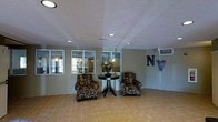 This single-story space presents a clean and functional lobby or common area designed to welcome visitors and occupants alike. The room features light-toned tile flooring that contributes to a bright, open atmosphere. Recessed lighting is installed throughout the textured white ceiling, providing even, consistent illumination across the space. Two patterned armchairs are arranged symmetrically around a dark wood pedestal table topped with a decorative floral arrangement, creating an inviting seating area at the center of the room. Large windows along one wall allow natural light to filter in while offering views of an adjacent area, adding depth to the space. A wall-mounted HVAC unit is positioned in the upper corner for climate control. A bold "NV" letter display mounted on the wall serves as a distinctive decorative feature, suggesting a branded or themed identity for the space. The overall layout is open and uncluttered, with neutral wall tones and simple finishes that give the area a clean, utilitarian yet welcoming character suited for a communal or reception environment.