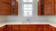Located at 12822 White Willow Dr in Plainfield, Illinois, this single-floor kitchen space is thoughtfully designed with both function and style in mind. The kitchen showcases rich, warm-toned wood cabinetry with sleek silver hardware, offering generous upper and lower storage throughout. Light-colored solid surface countertops provide ample workspace and contrast beautifully against the cabinetry's deep finish. A double undermount sink is positioned beneath a large double-hung window that fills the space with natural light and offers views of the surrounding neighborhood. A stainless steel top-freezer refrigerator adds a modern touch, while the decorative patterned tile flooring introduces a charming and distinctive character to the room. A standout architectural feature is the interior pass-through window, which opens the kitchen to an adjacent room showcasing elegant arched doorways and soaring ceilings — hinting at a grand, open layout beyond. This connection to the neighboring space enhances the overall sense of openness and flow. The clean white walls and bright natural light create a welcoming, airy ambiance throughout, making this kitchen a practical and visually appealing hub within the home.
