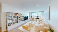 Located at The Breeze on East Broadway in Long Beach, NY, this well-appointed 2-bedroom, 3-bathroom residence offers a thoughtfully designed open-concept layout with light wood flooring and a clean, coastal aesthetic throughout. The combined living and kitchen area serves as the heart of the home, featuring a bright, airy living room with large modern windows that invite abundant natural light and views of the surrounding area. A built-in shelving unit provides stylish display and storage space alongside the entertainment area. The kitchen is outfitted with two-tone cabinetry — crisp white uppers paired with natural wood lowers — complemented by white countertops, stainless steel appliances including a French door refrigerator and range, and pendant lighting over the island. The spacious primary bedroom opens directly onto a private balcony and includes a generous walk-in closet with built-in shelving and hanging systems. The en-suite primary bathroom features a double vanity with backlit mirrors and a glass-enclosed shower with tile surround. The second bedroom is well-proportioned and filled with natural light. Additional highlights include a conveniently tucked-away stacked in-unit washer and dryer, two closets, and three full bathrooms across the residence.