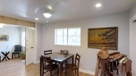 This single-story space presents a welcoming and functional interior highlighted by a casual dining area at its heart. The dining space accommodates a dark wood table paired with matching chairs, offering seating for up to four guests. A ceiling fan with a built-in light fixture provides both ventilation and ambient lighting, complemented by recessed lighting that ensures the area remains well-lit throughout the day. A horizontal window fitted with white blinds allows natural light to filter in, brightening the neutral gray-toned walls and light wood laminate flooring that runs throughout the space. A framed landscape painting mounted on the wall adds a warm, rustic character to the room, while decorative accents — including what appears to be a leather saddle stand — contribute to the space's distinct personality. A doorway visible along one wall suggests easy flow to adjacent rooms, enhancing the overall sense of openness. The clean white baseboards and simple architectural finishes give the space a tidy, understated aesthetic that is both practical and comfortable.