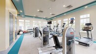 Located at 3500 Millenia Blvd in Orlando, Florida, this well-appointed fitness center offers a comprehensive exercise environment within a single, generously sized floor. The space is defined by its bright, modern aesthetic, featuring crisp white walls accented by bold teal coffered ceiling beams and warm yellow accent walls that create an energizing atmosphere.

Large multi-pane windows line the perimeter walls, flooding the room with natural light and offering views of the surrounding outdoor greenery. Recessed lighting throughout the ceiling ensures consistent illumination across all workout zones.

The fitness floor is organized into distinct functional areas. A dedicated cardio zone houses a row of treadmills, elliptical trainers, stationary bikes, and stair climbers, each equipped with individual digital display screens. A strength training section features a broad selection of cable and selectorized machines, including multi-functional cable systems and a variety of resistance equipment. A free weights area provides a full dumbbell rack alongside a flat bench, medicine balls, and stability balls for dynamic training.

The flooring combines neutral carpet with teal rubber mat zones that clearly delineate workout areas. Wall-mounted flat-screen televisions are positioned throughout for entertainment during workouts. The space is well-ventilated with visible HVAC systems integrated into the ceiling, ensuring a comfortable environment for all levels of exercise.