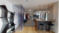 This thoughtfully designed single-floor coworking and community space offers a diverse range of environments tailored to productivity, socializing, and relaxation. The open layout flows seamlessly from zone to zone, anchored by a striking bar-height island clad in dark hexagonal tile with a warm wood countertop and pendant lighting — ideal for casual gatherings or informal work sessions. Light hardwood flooring transitions into plush carpet as the space moves deeper, with teal accent tiles adding playful pops of color throughout.

A glass-paneled coworking section provides a semi-private area for focused work, while a dedicated office features clean-lined desks, globe pendant lights, and floor-to-ceiling windows that draw in natural light and offer lush outdoor views. A comfortable lounge area with a large sectional sofa and oversized windows creates a welcoming retreat for relaxation.

Social areas include a cozy nook with a wall-mounted oversized Scrabble board, built-in booth seating with teal upholstery, and bold diagonal-stripe wall accents. Elsewhere, a dramatic deep-blue herringbone tile accent wall frames a seating area near a bold red-framed private room. A fireplace lounge with blue botanical wallcovering and warm pendant lighting rounds out this richly layered, community-centered space.