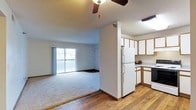 This single-floor residence offers a practical and comfortable open-concept layout that seamlessly connects the living area and kitchen. The living room is finished with plush carpeting and features a ceiling fan overhead, creating a relaxed and functional atmosphere. A large sliding glass door floods the space with natural light and opens onto a private balcony, offering views of the surrounding exterior. The transition between the living area and kitchen is defined by a partial dividing wall, providing a sense of separation while maintaining an open feel. The kitchen is outfitted with white cabinetry accented by wood-toned trim, offering ample storage space. A full-size refrigerator anchors the kitchen, which also features hardwood flooring that extends through the entryway and kitchen zones. The countertops provide generous workspace, and under-cabinet storage contributes to the kitchen's overall functionality. Textured ceilings are present throughout the space, adding a classic residential character. The combination of hardwood flooring in the kitchen and carpeting in the living area creates a clear and comfortable zoning between the two areas, making this a well-organized and inviting single-floor home.