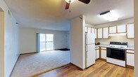 This single-floor residence offers a practical and comfortable open-concept layout that seamlessly connects the living area and kitchen. The living room is finished with plush carpeting and features a ceiling fan overhead, creating a relaxed and functional atmosphere. A large sliding glass door floods the space with natural light and opens onto a private balcony, offering views of the surrounding exterior. The transition between the living area and kitchen is defined by a partial dividing wall, providing a sense of separation while maintaining an open feel. The kitchen is outfitted with white cabinetry accented by wood-toned trim, offering ample storage space. A full-size refrigerator anchors the kitchen, which also features hardwood flooring that extends through the entryway and kitchen zones. The countertops provide generous workspace, and under-cabinet storage contributes to the kitchen's overall functionality. Textured ceilings are present throughout the space, adding a classic residential character. The combination of hardwood flooring in the kitchen and carpeting in the living area creates a clear and comfortable zoning between the two areas, making this a well-organized and inviting single-floor home.