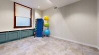 Located at 1501 Euclid Ave in Cleveland's vibrant Playhouse Square district, this fitness center offers a thoughtfully designed, single-floor layout comprising a generously sized exercise room, a laundry room, and a connecting hallway. The exercise room is the centerpiece of the space, featuring a striking multi-toned tile floor in shades of teal, blue, and cream that adds a modern, energetic feel. The room is outfitted with a comprehensive range of equipment, including multiple treadmills, elliptical machines, stationary bikes, cable and weight machines, free weights, medicine balls, stability balls, and yoga mats — catering to both cardio and strength training needs. Large windows with wood-trimmed frames allow natural light to fill the space, while recessed lighting and decorative ceiling details with dark trim accents provide a polished, finished look. Full-length mirrors along the walls enhance the sense of openness and allow for form checks during workouts. Teal-painted cabinetry adds a refined architectural touch throughout. The hallway connects the exercise room to the laundry facilities and building entry, featuring the same tile flooring and built-in lockers for convenient storage. A dedicated stretching and functional training area rounds out this well-appointed fitness facility.