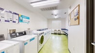 Located at 5100 W Alameda Ave in Denver, Colorado, this shared laundry facility offers a clean, efficiently organized space designed for practical, everyday use. The room is equipped with a row of top-loading washers and front-loading dryers, all branded with JETZnci signage indicating a coinless, app-based payment system — making the laundry process straightforward and convenient. Operating instructions are prominently posted along the wall above the machines in both English and Spanish, ensuring ease of use for all residents. A long, wall-mounted folding counter with a durable blue laminate surface runs the length of the opposite wall, providing ample space for sorting and folding laundry. Two folding chairs are positioned near a window fitted with horizontal blinds, offering a comfortable waiting area with natural light. The room features a vibrant green herringbone-patterned vinyl floor that adds a lively touch to the otherwise neutral white walls. Overhead fluorescent lighting ensures the space is well-lit throughout. A sturdy gray metal door provides secure access to the facility. The overall ambiance is tidy, functional, and well-maintained, reflecting a thoughtfully designed communal laundry environment.