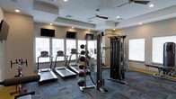 This fitness center offers a comprehensive workout environment designed to support a variety of exercise routines. The space is bright and open, benefiting from multiple windows along the walls that allow generous natural light to fill the room, complemented by recessed lighting and a ceiling fan that keeps the atmosphere comfortable during workouts.

The cardio section is outfitted with multiple treadmills and at least one elliptical machine, all positioned facing the windows to provide an engaging view during exercise. A medicine ball rack stocked with various weighted balls sits nearby, supporting functional and cross-training workouts. The strength training area features a cable machine and multi-functional rack, providing users with a range of resistance and weight training options.

Multiple flat-screen televisions are mounted along the walls at eye level, ensuring entertainment and motivation are always within view regardless of where you are training. The floor is covered in durable, patterned carpet tiles suited to a fitness environment. The ceiling features a tray design that adds a touch of architectural interest to the space. Overall, the fitness center is clean, well-lit, and thoughtfully arranged to maximize functionality and comfort for users of all fitness levels.