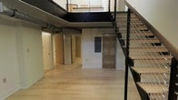 This two-story space showcases an industrial-modern aesthetic defined by its open layout and thoughtful architectural detailing. The lower level features light hardwood flooring that extends throughout, creating a bright and cohesive foundation. Exposed ductwork along the ceiling adds an authentic industrial character, complementing the raw, utilitarian feel of the space. White walls provide a clean contrast against the darker structural and mechanical elements visible overhead.

A striking open mezzanine level overlooks the main floor below, supported by dark-painted structural beams that contribute to the loft's bold visual identity. The upper level is accessed by a sleek staircase fitted with a contemporary cable railing system, combining safety with a minimalist design sensibility. The mezzanine's open railing allows sightlines to flow freely between floors, enhancing the sense of vertical volume throughout.

On the lower level, several doors lead to separate rooms or utility areas, offering functional versatility. The double-height ceiling amplifies the sense of spaciousness, while the interplay of natural and ambient light keeps the interior feeling open and airy. Overall, the space delivers a well-balanced blend of industrial character and modern refinement.