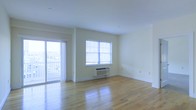 This single-floor space offers a clean, open layout highlighted by gleaming light hardwood floors that run throughout, reflecting abundant natural light and lending warmth to the interior. The main living area is bathed in brightness, courtesy of a large sliding glass door that opens to a private balcony, providing outdoor views and excellent ventilation. A secondary window on the adjacent wall further enhances the flow of natural light, keeping the space feeling airy and expansive. The walls are finished in a soft, neutral tone that complements the light flooring and contributes to the overall bright and welcoming ambiance. A wall-mounted PTAC heating and cooling unit beneath the window ensures year-round climate control. The space features clean white baseboards and smooth ceilings with recessed lighting, adding to its polished, move-in-ready appeal. The open plan offers flexible furniture arrangement possibilities, making it well-suited for a variety of uses. Overall, the space presents a straightforward, functional design with an emphasis on natural light, clean finishes, and easy indoor-outdoor connectivity through the balcony access.