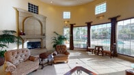 This single-story community clubhouse at 26900 Winchester Creek Ave in Murrieta, California, presents a refined Mediterranean-inspired aesthetic from the moment you arrive. The entrance is marked by a grand arched doorway with solid wood double doors set beneath a barrel-vaulted ceiling and an ornate pendant light, establishing an immediately welcoming and upscale tone.

Inside, the main lounge area features soaring ceilings with clerestory windows that flood the space with natural light. Large floor-to-ceiling windows and glass doors offer direct views of the outdoor pool area, seamlessly connecting the interior to the exterior. The room is anchored by a stately stone fireplace with a classic arched mantel, complemented by plush seating arrangements on soft carpeting. Decorative draperies and plantation shutters add warmth and elegance throughout.

An adjacent lounge and reception area showcases polished travertine tile flooring, high ceilings, recessed lighting, and built-in niche accents with decorative mirrors and ornamental furnishings. Lush tropical plants are positioned throughout, enhancing the inviting ambiance.

A separate conference room offers a more focused setting, furnished with a wooden conference table and executive chairs, wood-shuttered windows, and tasteful framed artwork — ideal for meetings and community gatherings. The overall design blends comfort and sophistication, making this clubhouse a distinguished amenity space.
