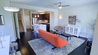 This two-story space makes a strong visual impression through its open-concept layout, where the living area and kitchen flow naturally into one another. Dark hardwood flooring runs throughout the main level, providing a rich contrast against the crisp white walls and light-toned ceiling. A ceiling fan with a brushed nickel finish anchors the living area overhead, contributing both function and a sleek aesthetic.

The kitchen is well-appointed with dark wood cabinetry featuring a bold grain pattern, stainless steel appliances including a refrigerator, microwave, and range, and a peninsula counter with bar-height seating. Pendant lighting hangs above the peninsula, adding warmth to the cooking and dining zone.

The living area accommodates a modern furniture arrangement, with a plush area rug defining the seating space. A staircase along the far wall leads to the second floor, hinting at additional living space above. Wall-mounted lighting and curated décor, including colorful artwork, add character throughout. Overall, the space balances contemporary design with a comfortable, livable atmosphere.