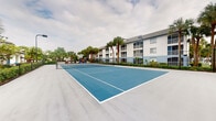 This outdoor recreational space features a well-maintained tennis court with a striking blue playing surface, clearly marked white boundary lines, and a regulation-height net positioned at center court. The court is finished in a smooth, hard-surface material designed for consistent play and durability. Surrounding the court, tall palm trees provide a lush, tropical backdrop that adds to the open, airy atmosphere of the space. The court is set within a multi-story residential community, with a white and blue-accented building visible in the background, offering balconied units that overlook the recreational area. A playground area can also be seen adjacent to the court, indicating a broader outdoor amenity zone designed for community use. The court benefits from open-air exposure, allowing for natural ventilation and an unobstructed view of the sky. The surrounding landscape is neatly maintained with low-lying shrubs and greenery forming a natural perimeter. The overall setting conveys a clean, well-kept community environment that prioritizes outdoor activity and leisure, making this tennis court a central feature of the property's recreational offerings.