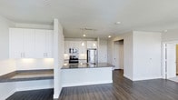 Located at The Colony, this single-floor residence offers a clean, modern aesthetic defined by an open-concept layout and thoughtfully selected finishes throughout. The kitchen serves as a central focal point, featuring crisp white cabinetry that extends to the ceiling, a classic white subway tile backsplash, and dark granite countertops that provide a striking contrast. A spacious kitchen island with matching countertop offers additional prep space and doubles as a casual dining or gathering area, illuminated by elegant pendant lights hanging overhead. Stainless steel appliances, including a refrigerator, range, and microwave, complete the contemporary kitchen setup. Recessed and track lighting ensure the space is well-lit throughout. Rich, dark hardwood-style flooring runs seamlessly across the open living area, grounding the bright white palette and adding warmth to the overall ambiance. A dedicated entryway with a closet provides practical storage upon arrival. The overall design balances functionality with modern style, creating a cohesive and inviting living environment with a bright, airy feel courtesy of the neutral color scheme and open floor plan.
