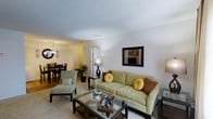 This single-floor residence offers a cohesive and inviting layout designed for comfortable everyday living. The living area serves as the heart of the home, centered around a plush sage-green sofa accented with gold and brown throw pillows, complemented by a glass-topped coffee table and a coordinating accent chair. Neutral carpeting and white walls create a bright, clean backdrop that enhances the warm ambiance of the space. A stylish table lamp adds a soft, welcoming glow to the room. The living area flows seamlessly into the dining space, which features a dark wood dining table and chairs beneath an elegant chandelier, lending a touch of sophistication to the open-plan arrangement. Lush indoor greenery adds a natural element and visual interest to the transition between rooms. A textured ceiling adds subtle architectural character throughout. The layout connects efficiently to additional areas of the home via a well-lit hallway, suggesting a practical and well-organized floor plan. Overall, the space exudes a warm, neutral aesthetic with thoughtfully coordinated furnishings, making it a comfortable and functional single-level home.