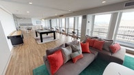 This contemporary single-floor lounge space is thoughtfully designed to accommodate a variety of social and recreational activities. The open floor plan allows for a seamless flow between distinct functional zones, all unified by warm wood-tone flooring and a clean, modern aesthetic featuring white and grey tones accented with pops of teal, coral, and yellow.

The main gathering area centers around generous sectional sofas and a teal area rug, paired with a wall-mounted large-screen television and a walnut media console. A bold abstract artwork adorns one accent wall, complemented by wall-mounted lighting. Adjacent to this is a recreational zone housing a full-size pool table, surrounded by bar-height seating positioned along the perimeter windows.

A casual dining nook features a round white pedestal table with modern chairs, while a sleek kitchenette with grey quartz countertops, white cabinetry, an under-counter beverage fridge, and a utility sink provides functional support for gatherings.

Floor-to-ceiling windows line the exterior walls throughout, flooding the space with abundant natural light and offering sweeping urban skyline views. The overall ambiance is polished yet inviting, making it well-suited for both relaxed social interaction and organized events.