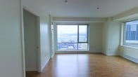 This single-floor space is designed with a clean, modern aesthetic that emphasizes openness and natural light. The main living area is anchored by a large picture window framed in dark trim, which floods the room with daylight and offers elevated views of the surrounding urban cityscape, including rooftops and city infrastructure beyond. The window's generous proportions make it a standout architectural feature, visually expanding the space and connecting the interior to the dynamic city environment outside.

The flooring throughout the living area consists of warm-toned hardwood, which adds a sense of warmth and continuity to the otherwise minimalist interior. Crisp white walls and white baseboards complement the light wood floors, creating a neutral, versatile backdrop well-suited for a variety of interior styles. The ceiling is smooth and white, with recessed fixtures providing supplemental lighting alongside the abundant natural light.

A doorway along one wall suggests additional rooms or storage beyond the main living area. The overall layout feels open and uncluttered, with clean lines throughout. The elevated position of the unit, as evidenced by the rooftop-level views, enhances the sense of privacy and urban connection simultaneously.