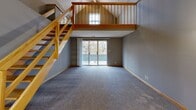 Located at 3100 S 72nd Street in Lincoln, Nebraska, this one-bedroom, one-bathroom loft-style unit offers a thoughtfully designed layout with standout architectural character. The main living area immediately impresses with soaring vaulted ceilings and a striking open wood staircase leading to an upper loft-style landing. A sliding glass door floods the carpeted living space with natural light and provides views of mature surrounding trees, enhancing the sense of openness. The kitchen features gray shaker-style cabinetry, light quartz countertops, hexagonal tile flooring, and stainless steel appliances including a range and over-the-range microwave. A convenient pass-through window connects the kitchen to the entry area, which also includes in-unit washer and dryer hookups. The spacious bedroom is carpeted, finished with warm wood baseboards, and includes a ceiling fan, natural light from multiple windows, and generous closet storage. The full bathroom is clean and well-appointed, offering a combined tub and shower, a vanity with storage, and matching hexagonal tile flooring. The upper loft landing, bathed in light from a transom window, adds flexible additional space and visual drama to this uniquely designed home.