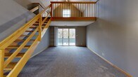 Located at 3100 S 72nd Street in Lincoln, Nebraska, this one-bedroom, one-bathroom loft-style unit offers a thoughtfully designed layout with standout architectural character. The main living area immediately impresses with soaring vaulted ceilings and a striking open wood staircase leading to an upper loft-style landing. A sliding glass door floods the carpeted living space with natural light and provides views of mature surrounding trees, enhancing the sense of openness. The kitchen features gray shaker-style cabinetry, light quartz countertops, hexagonal tile flooring, and stainless steel appliances including a range and over-the-range microwave. A convenient pass-through window connects the kitchen to the entry area, which also includes in-unit washer and dryer hookups. The spacious bedroom is carpeted, finished with warm wood baseboards, and includes a ceiling fan, natural light from multiple windows, and generous closet storage. The full bathroom is clean and well-appointed, offering a combined tub and shower, a vanity with storage, and matching hexagonal tile flooring. The upper loft landing, bathed in light from a transom window, adds flexible additional space and visual drama to this uniquely designed home.